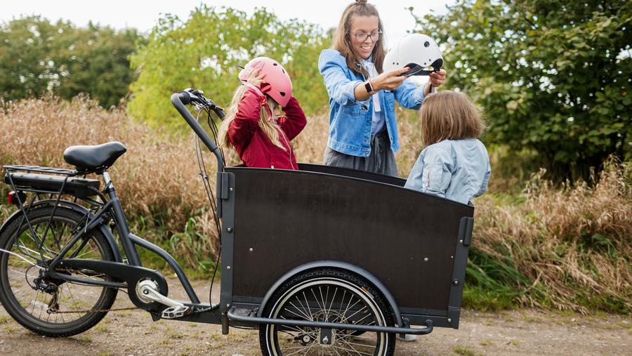 Mamma med två barn i lådcykel. Mamman klär på cykelhjälmar på barnen. 